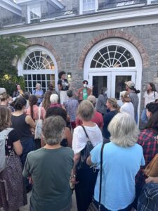 A crowd of people listen to speakers in front of the doors of an old stone building.